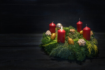 Light in the dark on first advent, natural green wreath with red candles, one is burning, Christmas decoration and cookies, dark wooden background, copy space