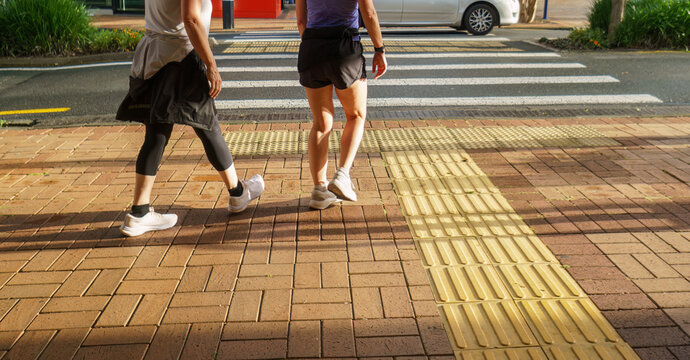 Two Women Walking To The Pedestrian Crossing. A Car Driving Past The Other Side Of The Crossing. Auckland.