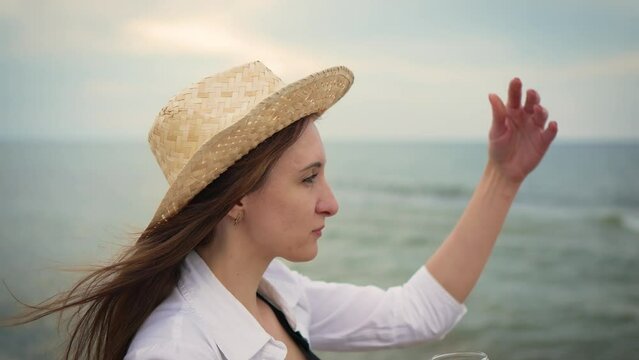 Portrait brunette Woman drinking wine wearing in straw hat and white shirt at sea beach. Alcohol drinking, enjoying life, active rest at seashore