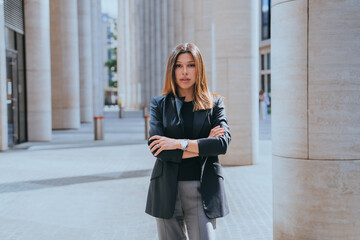 Purposeful young woman in business suit standing against huge columns looks at camera folding hands...