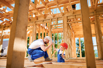Father with toddler son building wooden frame house. Happy worker giving high five to smiling kid...