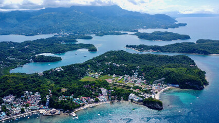Top view of a small town on the shore of a lagoon on a tropical island. Aerial view of a coastal town among green forest on the shore of the blue sea. Sabang, Puerto Galera, Philippines. - obrazy, fototapety, plakaty