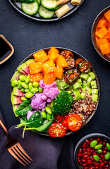 Vegan buddha bowl with pumpkin, quinoa, tomatoes, spinach, celery, radish, soybeans, edamame, tofu, cauliflower, broccoli and seeds, black table background, top view. Autumn or winter healthy food