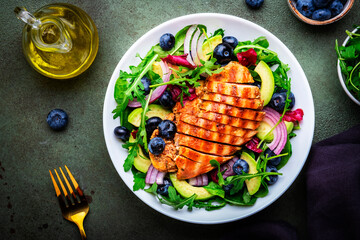 Fresh salad with blueberries, grilled chicken breast, avocado, red onion, walnuts and arugula on green table background, top view