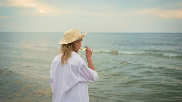 Blonde woman, wearing straw hat and white shirt, enjoying wine at sea beach, thoughtfully looking at waves. Woman at ocean with alcohol, champagne or drink for celebration, vacation party