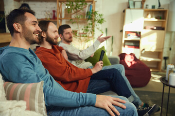 group of friends, men talking and having fun while watching TV sitting on sofa in living room at...