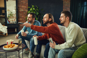 Happy and cheerful young men with alcohol drinks and snacks watching football game sitting on coach in living room at home. - obrazy, fototapety, plakaty