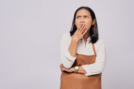 Surprised young Asian woman barista barman employee in brown apron working in coffee shop, posing covered mouth with hand, looking aside isolated on white background. Small business startup concept