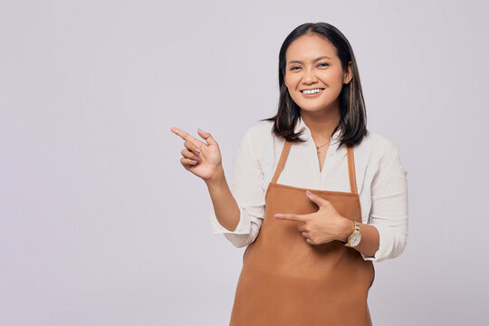 smiling young Asian woman barista barman employee wearing a brown apron working in coffee shop, pointing finger on advertising area isolated on white background. Small business startup concept