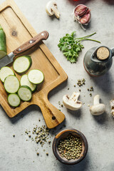 Fototapeta na wymiar Healthy cooking concept. Zucchini slices on cutting board with knife, herbs, mushrooms and utensils on kitchen table, top view