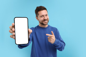 Handsome man showing smartphone in hand and pointing at it on light blue background, selective...