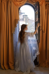 A young girl in a white vintage dress from the early 20th century looks out the window of her castle