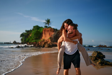 Fototapeta premium Loving couple enjoys honeymoon on exotic beach. Man carries woman piggyback, strolling along sea. Carefree lovers embrace, walk on sand near ocean, cliff. Romantic getaway, unity, joy, love.