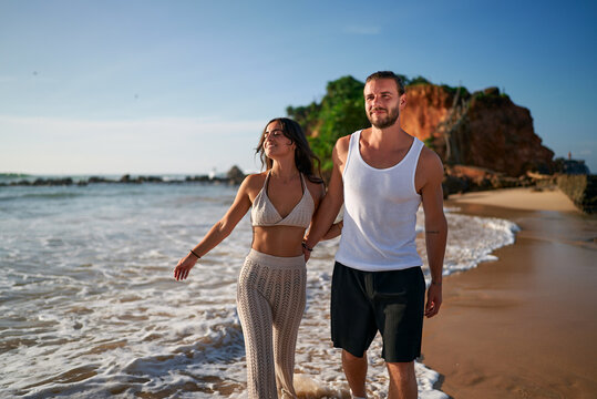 Romantic couple strolls on beach at sunset. Newlyweds on honeymoon enjoy sea, sand near cliffs. Man in tank top, woman in skirt walking barefoot, holding hands. Love, travel, vacation concept captured