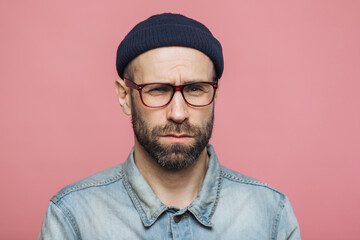 Confused man with a beard and glasses wearing a beanie and denim shirt on a pink background