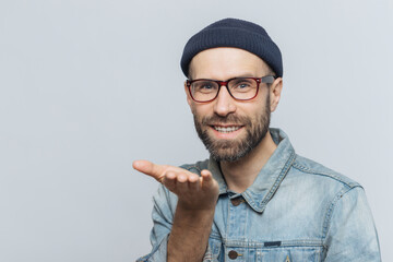 Smiling bearded man in glasses and beanie presenting something on his open hand, against a gray background.