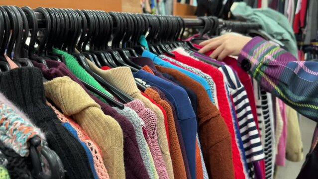 Woman hand chooses clothes in a second-hand shop. Thrift store hangers with colorful winter sweaters. Fast fashion industry waste reduce and sustainability concept. Selective focus on foreground.