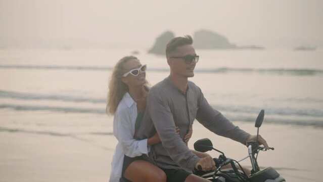 Elegant couple ride motorcycle along sea beach on sunrise. Intraframe installation from bike to people. Tourists drive rented motorbike on ocean coast. Woman hugs man, spreads arms riding scooter.