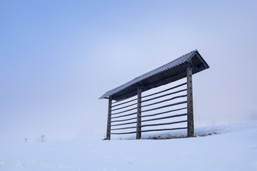 A hay stand on a snow-covered field against a light sky in a haze in the Alpine foothills of Slovenia