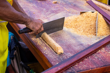 Hand Cutting a Roll of Smashed Caramelized Peanut by A Cleaver into Pieces on a Wooden Table Besides Crushed Peanut Heap