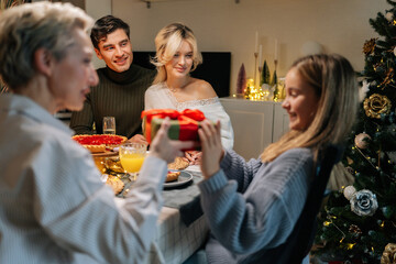 Portrait of loving grandmother giving festive box with Christmas present to adorable granddaughter sitting at dinner feast table during family party. Concept of home festive atmosphere.