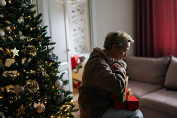 Obraz na płótnie Canvas Side view of happy middle-aged woman holding big pile of wrapped gifts, sitting by decorated Christmas tree, smiling looking on presents. Concept of home festive atmosphere.