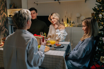 Back view of unrecognizable surprised woman receiving Christmas gift from loving family at home on xmas eve. Loving daughter giving present to mother on holidays. Concept of festive atmosphere.