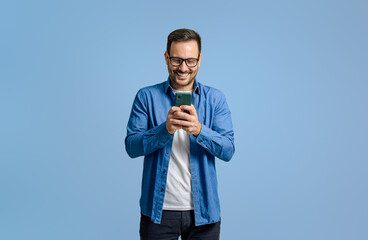 Male freelancer smiling and using social media over smart phone while standing on blue background