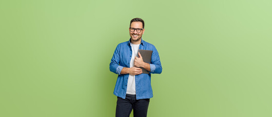 Portrait of handsome entrepreneur holding digital tablet and smiling at camera on green background - obrazy, fototapety, plakaty