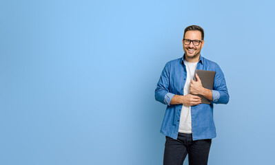 Confident young businessman holding digital tablet and smiling at camera against blue background - obrazy, fototapety, plakaty