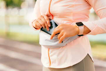 Middle aged woman hands are putting in waist pouch bag her smart phone outdoors.