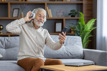 Senior man sitting on sofa at home and resting. Wearing white headphones, he listens to music from...