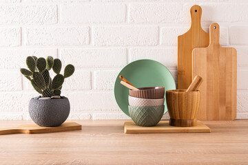 Set of ceramic bowls and cutting boards on wooden light countertop in modern kitchen with potted opuntia, cactus. Front view. minimalism.