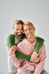 joyful bearded gay man embracing stylish boyfriend smiling with closed eyes on grey backdrop