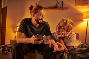happy bearded gay man browsing internet on mobile phone near smiling boyfriend in bedroom at night