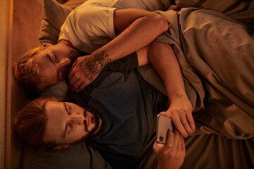 top view of unfaithful bearded gay man chatting on smartphone near sleeping boyfriend at night