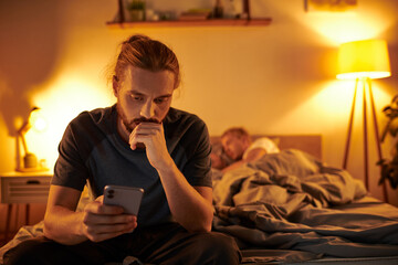 disloyal bearded gay man browsing internet on smartphone near partner sleeping at night in bedroom - obrazy, fototapety, plakaty