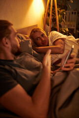 disloyal gay man browsing date app on smartphone near discouraged boyfriend at night in bedroom