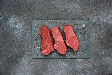 Three raw beef steaks on a black slate board on a dark concrete background. Steak ingredients, raw...
