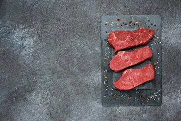 Three raw beef steaks on a black slate board on a dark concrete background. Steak ingredients, raw...