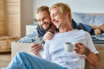 cheerful bearded gay man smiling near boyfriend with coffee cup using mobile phone in bedroom