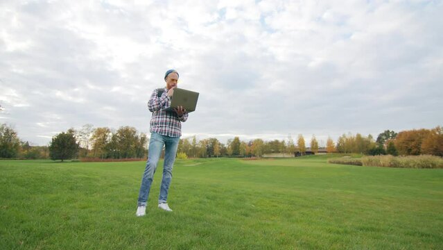 Vast territory with green lawn, distant multicolored trees in autumn. Young male thoughtfully touching his beard, thinking of solution, looking into laptop. High quality 4k footage