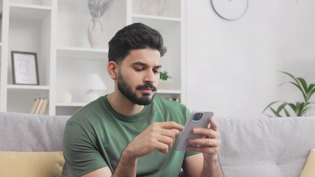 Thoughtful millennial guy seriously looking in distance while sitting on sofa and holding smartphone. Hindu man in green t-shirt translating view on mobile screen and beaming in smile.