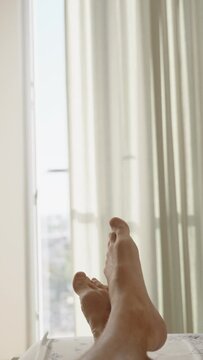 Vertical video. Top-down view of a white room illuminated by bright sunlight through a panoramic window and a sliver through the curtain. First-person view of male legs on the bed.