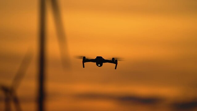Drone inspecting Work of wind farm with windmill turbines park, quadrocopter uses for study advantages and drawbacks of electric station at incredible multi colored cloudy sunset. Wind farm generating