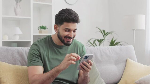 Handsome indian man sitting on couch, opening mouth and making big eyes while looking on smartphone screen with surprised facial expression. Unexpected news and reaction concept.