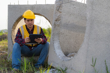 Young Asian civil engineer or construction supervisor wearing a helmet looks away and smiles while inspecting a construction site.