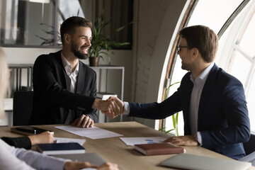 Confident cheerful boss and project manager man in formal suits shaking hands over meeting table, smiling, discussing deal, contract, investment, project success, starting partnership, cooperation