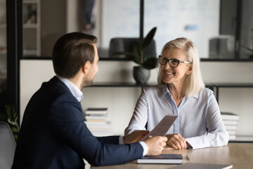 Two happy older and younger business partners speaking at meeting table, discussing paper contract. Employer and candidate talking on job interview, negotiating on resume - obrazy, fototapety, plakaty