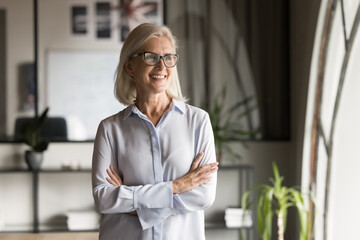 Happy pensive elder business leader woman office portrait. Confident senior businesswoman in elegant glasses, mature professional with arms crossed looking away with toothy smile